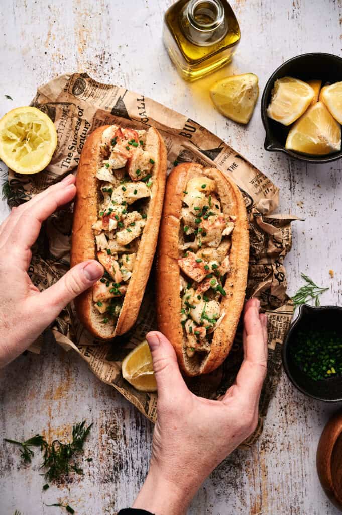 Two hands hold classic lobster rolls in buns on newspaper, surrounded by lemon wedges, herbs, chives, and a bottle of olive oil on a rustic surface.