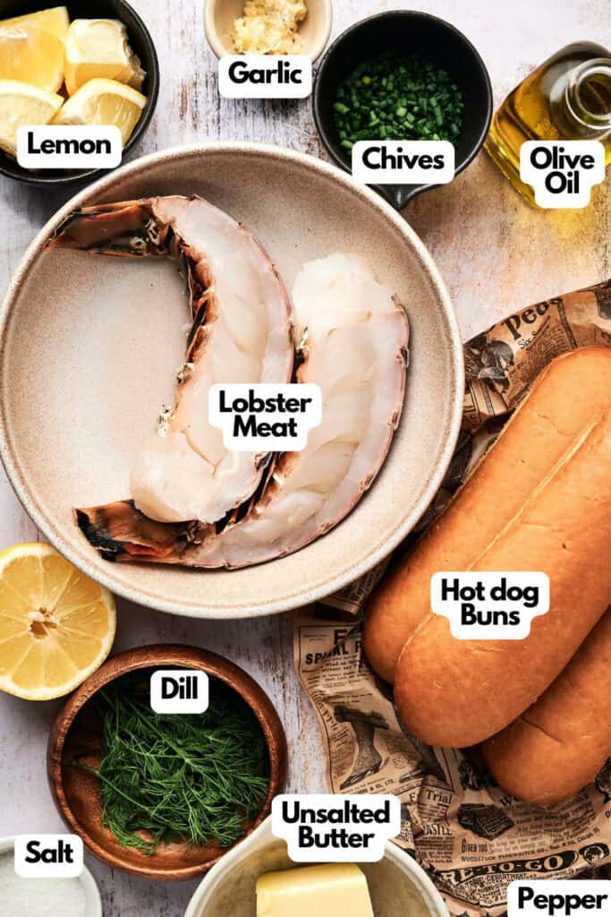 A top-down view of lobster roll ingredients, including lobster meat, hot dog buns, lemon, garlic, chives, olive oil, dill, unsalted butter, salt, and pepper.