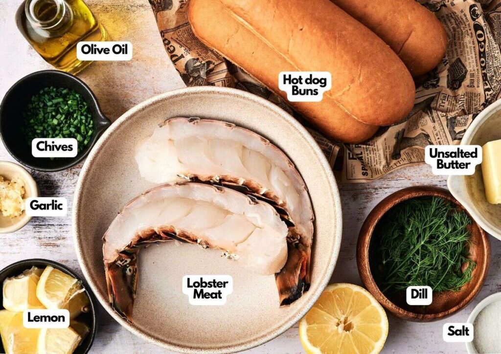 Top-down view of lobster meat, hot dog buns, olive oil, unsalted butter, chives, garlic, dill, salt, and lemon arranged on a table and labeled—everything you need for the perfect lobster roll.