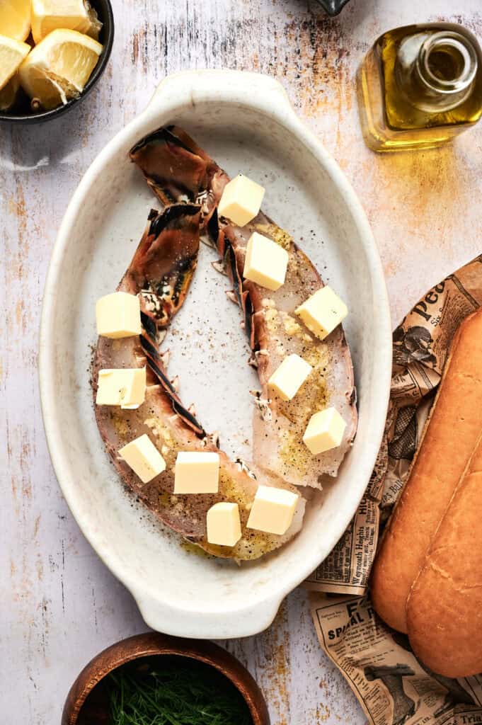 Two uncooked lobster tails in a white oval dish, topped with butter cubes and pepper, surrounded by lemon wedges, olive oil, bread, and herbs—perfect ingredients for crafting a classic lobster roll at home.