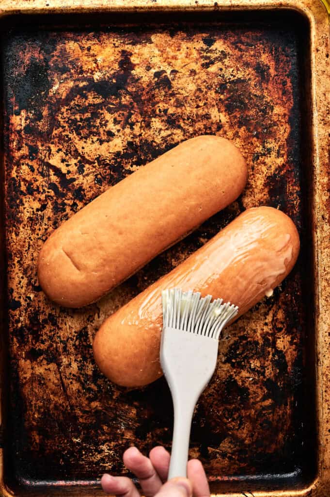 A hand uses a pastry brush to apply melted butter to two hoagie rolls—perfect for assembling a classic lobster roll—on a worn baking sheet.