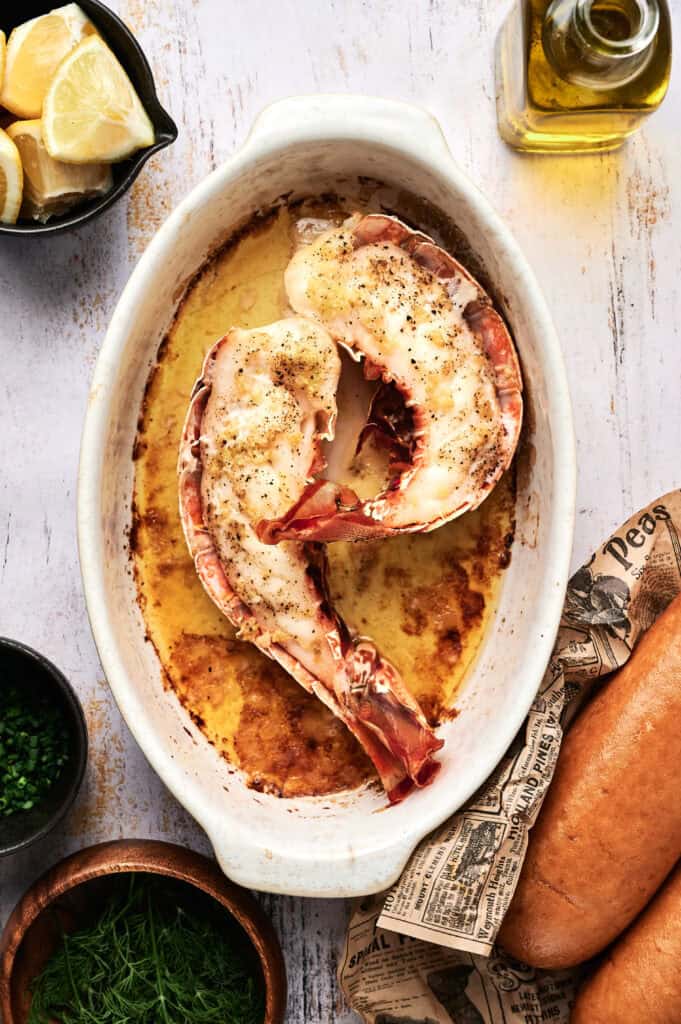Two baked lobster tails in a white oval dish, surrounded by lemon wedges, olive oil, fresh herbs, and a baguette&mdash;perfect for pairing with a classic lobster roll on a rustic surface.