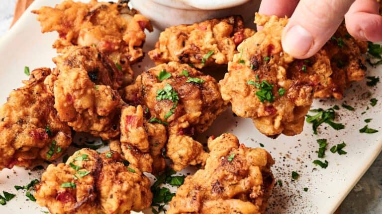 A hand reaches for a piece of golden-brown fried chicken on a white plate, garnished with chopped herbs and served with a dipping sauce.