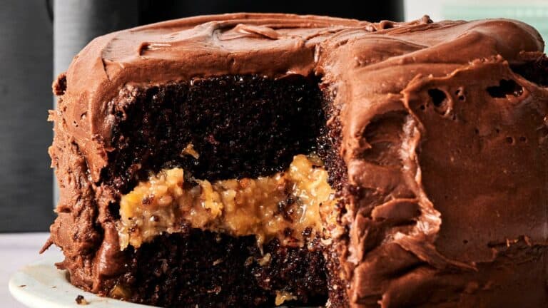 A close-up of a chocolate cake with chocolate frosting and a visible filling layer of coconut and pecans, with one large slice removed.