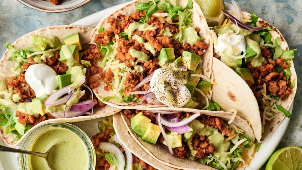 Three soft tacos filled with seasoned ground meat, shredded lettuce, diced avocado, red onion, shredded cheese, and topped with sour cream and green sauce sit on a plate.