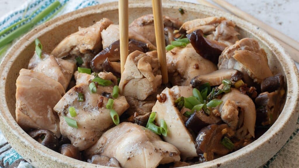 A bowl of cooked chicken pieces and mushrooms garnished with chopped green onions, with chopsticks picking up a piece.