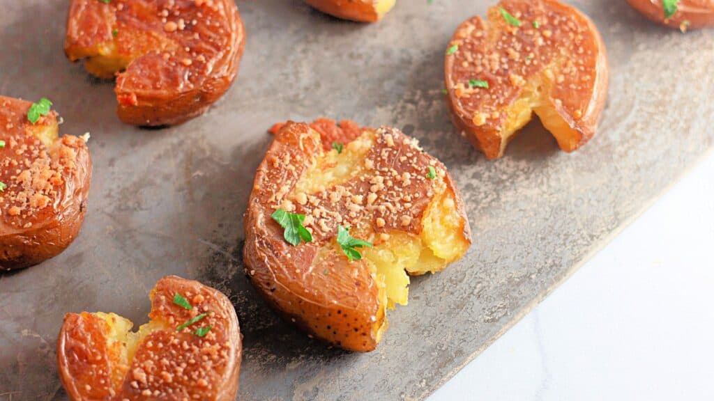 Several smashed, roasted baby potatoes garnished with herbs and grated cheese are arranged on a baking sheet.