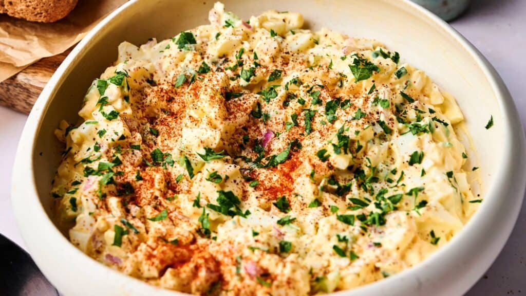 A bowl of creamy egg salad topped with chopped parsley and a sprinkle of paprika.
