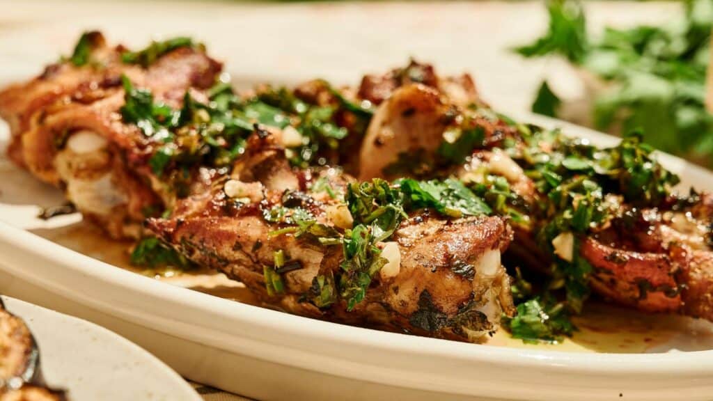 Grilled lamb chops topped with a herb and garlic mixture are arranged on a white platter with fresh greens in the background.