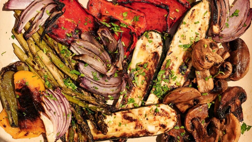 A platter of grilled vegetables including red onions, bell peppers, asparagus, zucchini, and mushrooms, garnished with chopped herbs.
