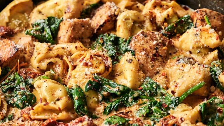 Close-up of creamy tortellini pasta with spinach, grilled chicken slices, and grated cheese, garnished with red pepper flakes.