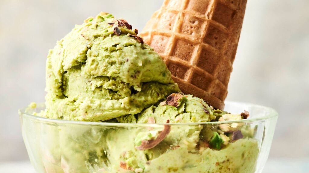 A glass bowl holds two scoops of pistachio ice cream topped with crushed pistachios, with a waffle cone placed upside down in the bowl.