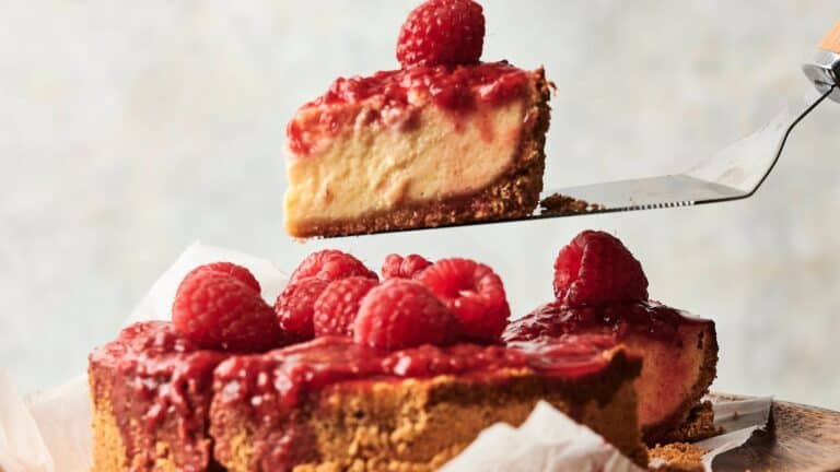 A slice of raspberry cheesecake is being lifted from a whole cheesecake topped with raspberry sauce and fresh raspberries.