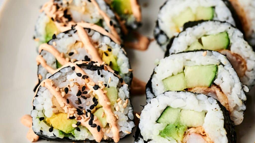 Close-up of sushi rolls filled with avocado, cucumber, and rice, topped with spicy mayo sauce and sprinkled with black and white sesame seeds on a beige plate.