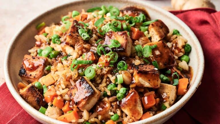 A bowl of fried rice with grilled tofu cubes, diced carrots, peas, chopped green onions, and a drizzle of dark sauce, served on a red cloth.