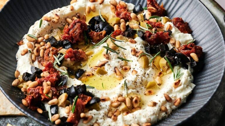 A bowl of whipped feta topped with pine nuts, black olives, sun-dried tomatoes, rosemary, olive oil, and ground pepper.