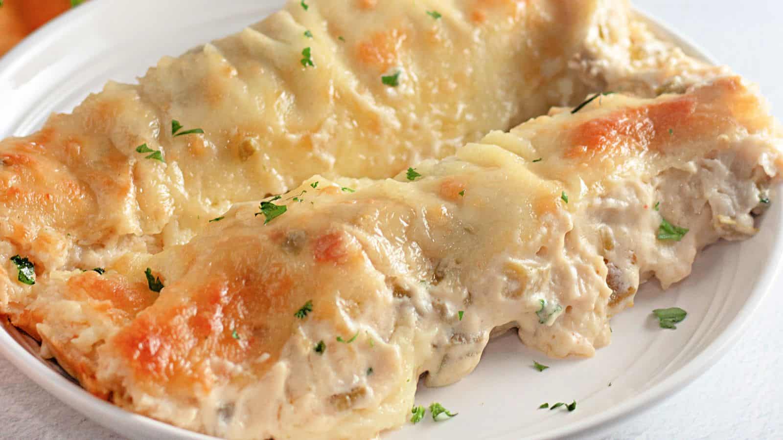 Two creamy, cheese-topped chicken enchiladas are served on a white plate, garnished with small pieces of parsley.