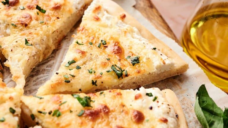 Close-up of sliced white pizza with melted cheese, herbs, and a golden crust on parchment paper, next to a glass of olive oil.