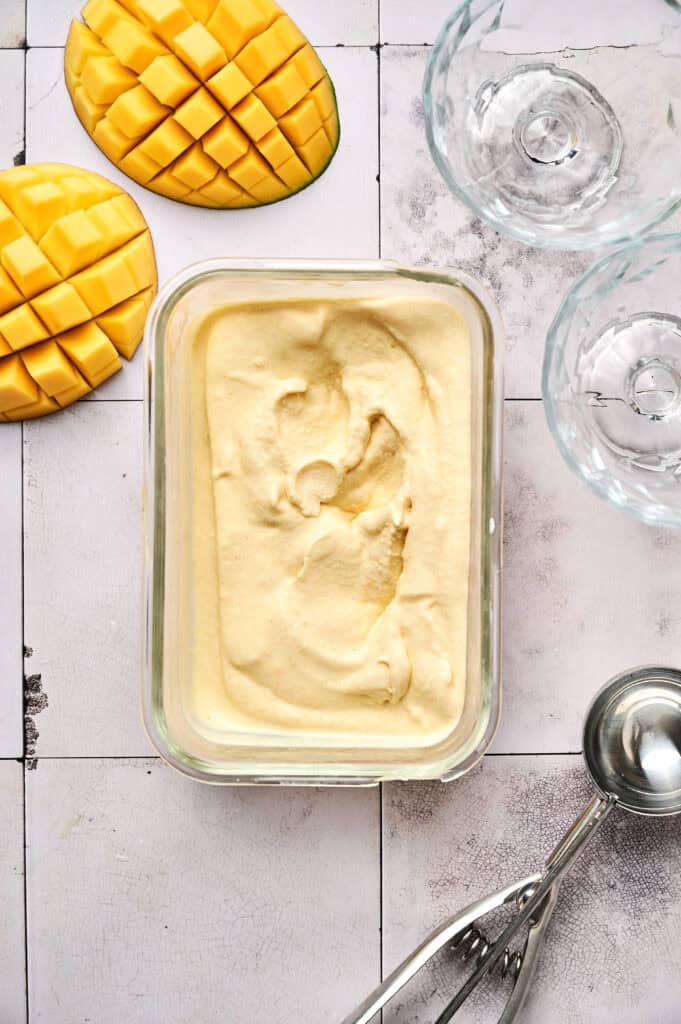 A rectangular glass container filled with mango ice cream sits on a tiled surface next to fresh mango halves, glass bowls, and a metal scoop, highlighting the creamy texture of this delicious mango ice cream dessert.