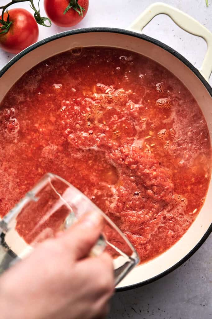 A hand pours water into a pot of crushed tomatoes on a light surface as whole tomatoes and ingredients for mexican rice are visible nearby.