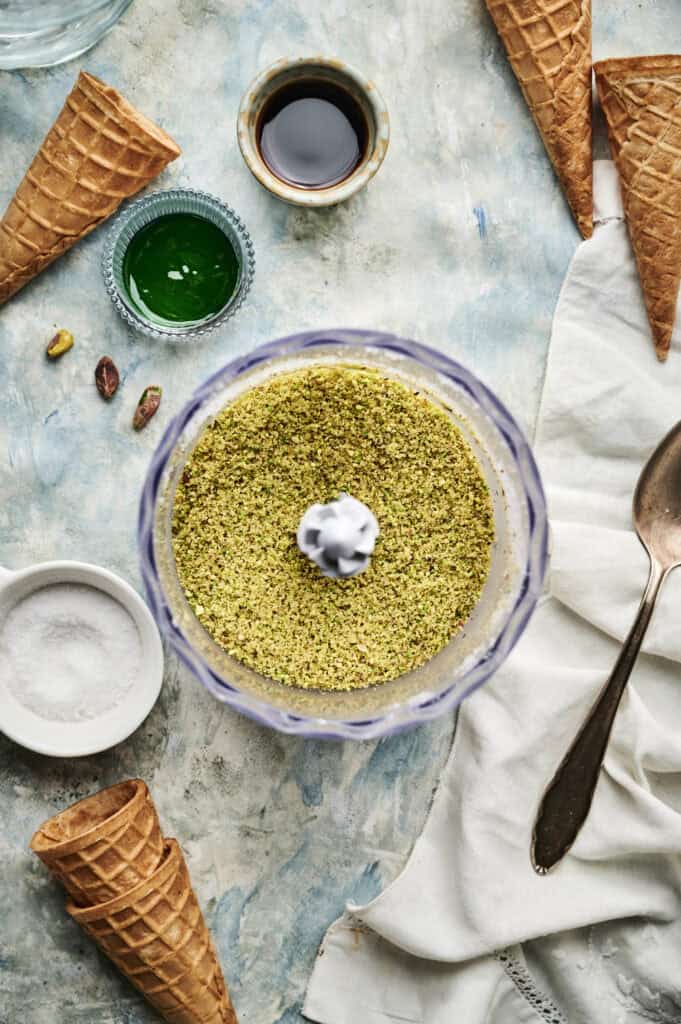 Ground pistachios in a food processor bowl are surrounded by ice cream cones, a spoon, salt, green liquid, and a small dish of dark sauce—perfect ingredients for making Pistachio Ice Cream on a textured surface.