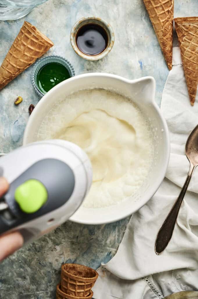 A hand holds a mixer blending a white mixture in a bowl, surrounded by empty waffle cones, a spoon, and small bowls of ingredients—perfect for creating homemade pistachio ice cream on a textured surface.