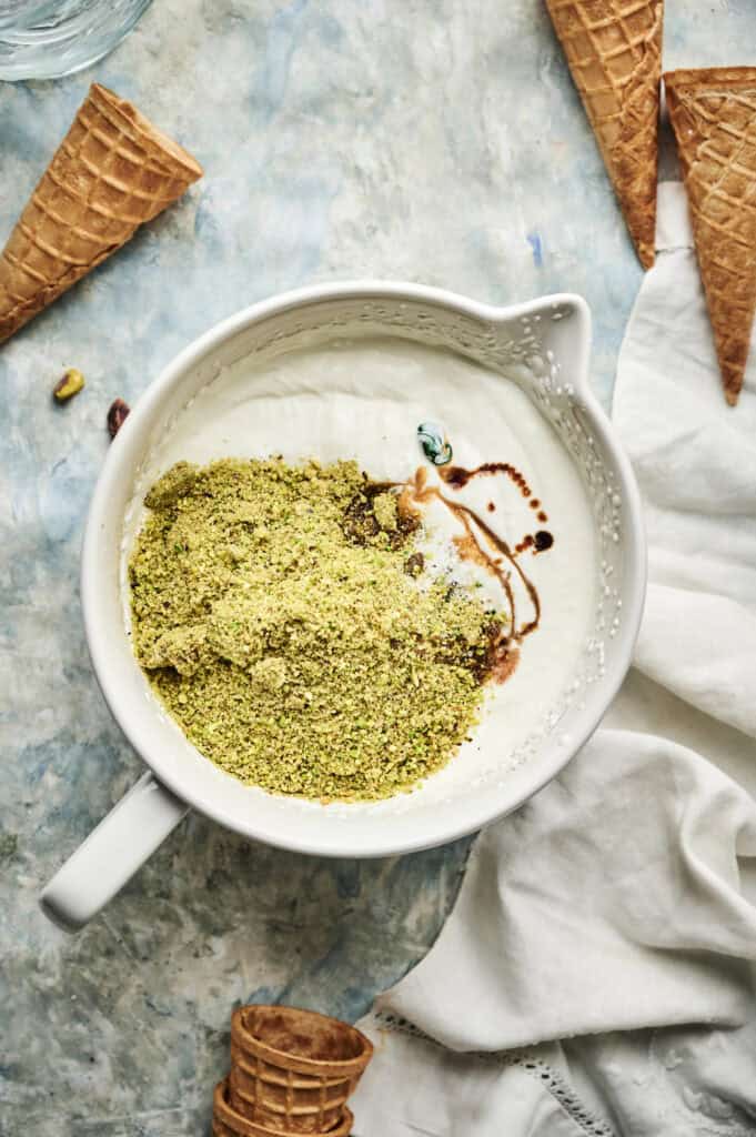 A white bowl holds the makings of Pistachio Ice Cream—ground pistachios, yogurt, and vanilla extract—surrounded by empty waffle cones and a white napkin on a textured surface.