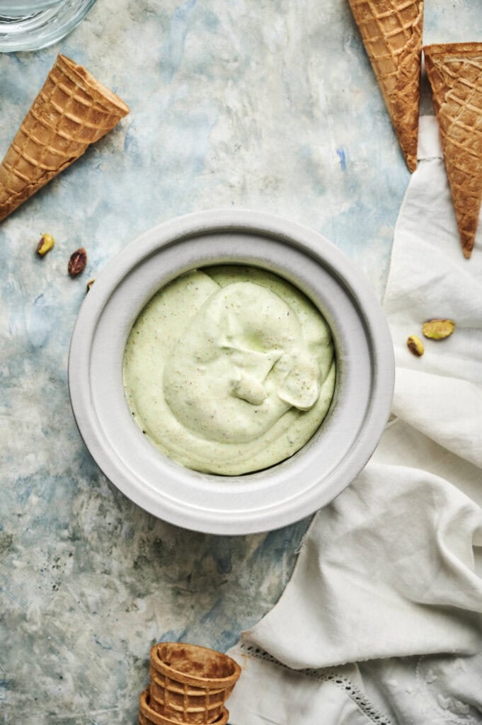A bowl of pale green Pistachio Ice Cream sits beside empty waffle cones, a white cloth, and a few scattered pistachios on a light blue textured surface.