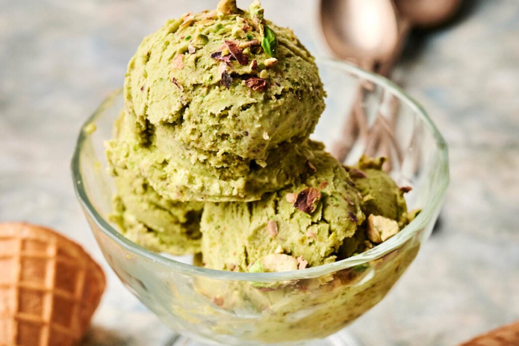 Three scoops of creamy Pistachio Ice Cream are served in a glass bowl, topped with a generous sprinkle of chopped pistachios. A waffle cone and spoons are visible in the background, adding to the inviting scene.