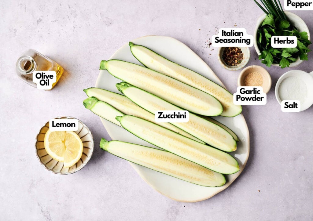 Four halved zucchinis on a plate with labeled ingredients around them: olive oil, lemon slices, Italian seasoning, garlic powder, salt, pepper, and fresh herbs&mdash;everything you need for flavorful baked zucchini.