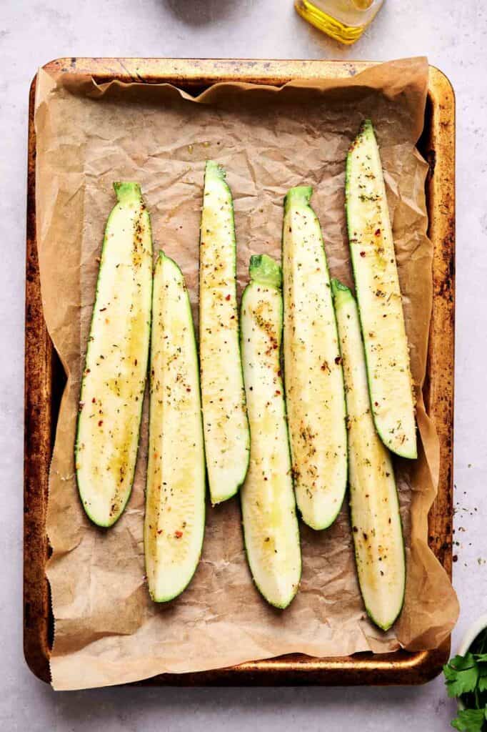Sliced zucchini halves arranged on a parchment-lined baking sheet, seasoned with spices and drizzled with oil, ready to become delicious baked zucchini.