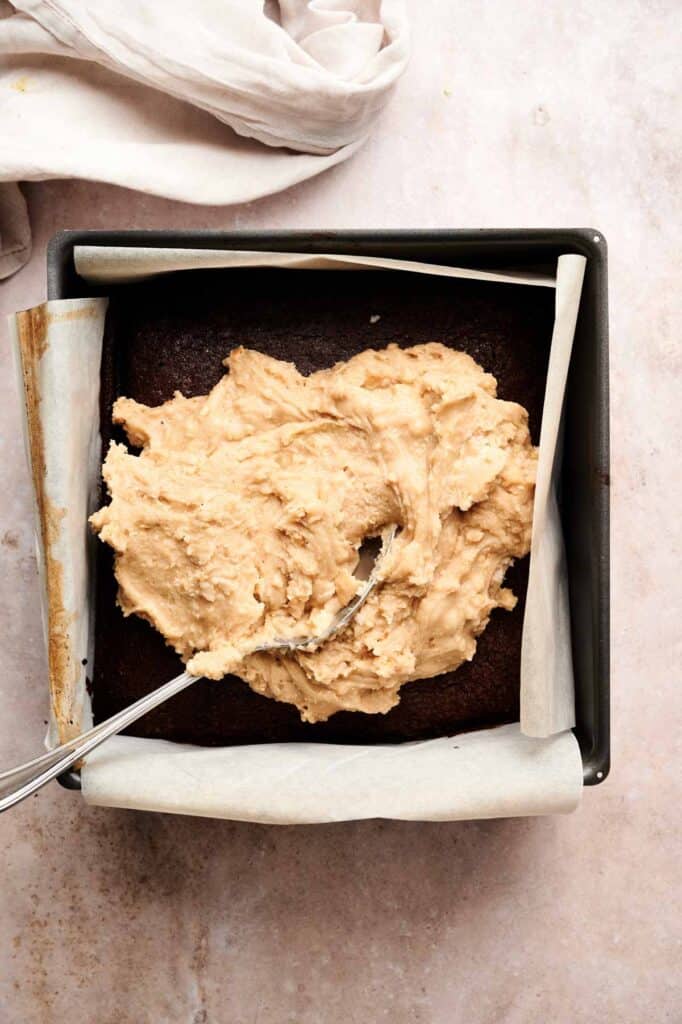 A square baking pan with a chocolate cake base lined with parchment paper, topped with a thick layer of beige frosting being spread with a spoon&mdash;perfect for making decadent buckeye brownies.