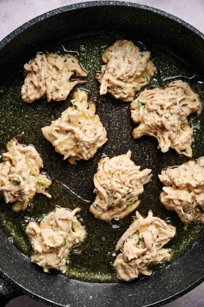 Raw chicken fritter batter portions with visible shredded vegetables are arranged in a frying pan with oil, ready to be cooked.