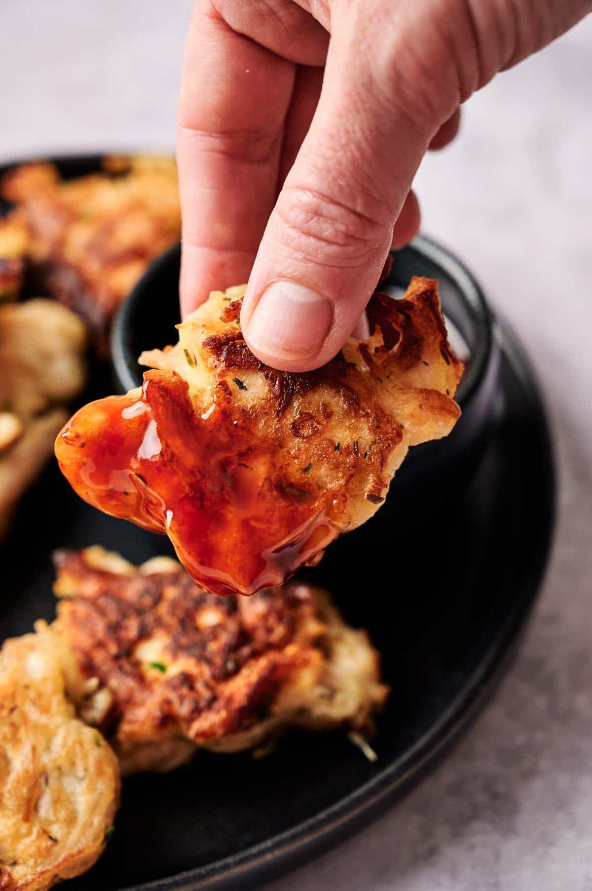 A hand dips a piece of crispy chicken fritter into a small bowl of red sauce on a black plate.