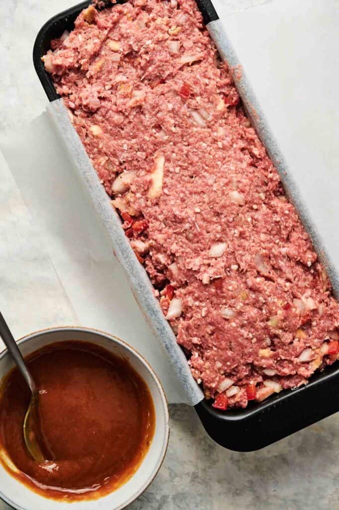 Uncooked Cracker Barrel Meatloaf mixture in a parchment-lined loaf pan next to a bowl of brown sauce with a spoon.
