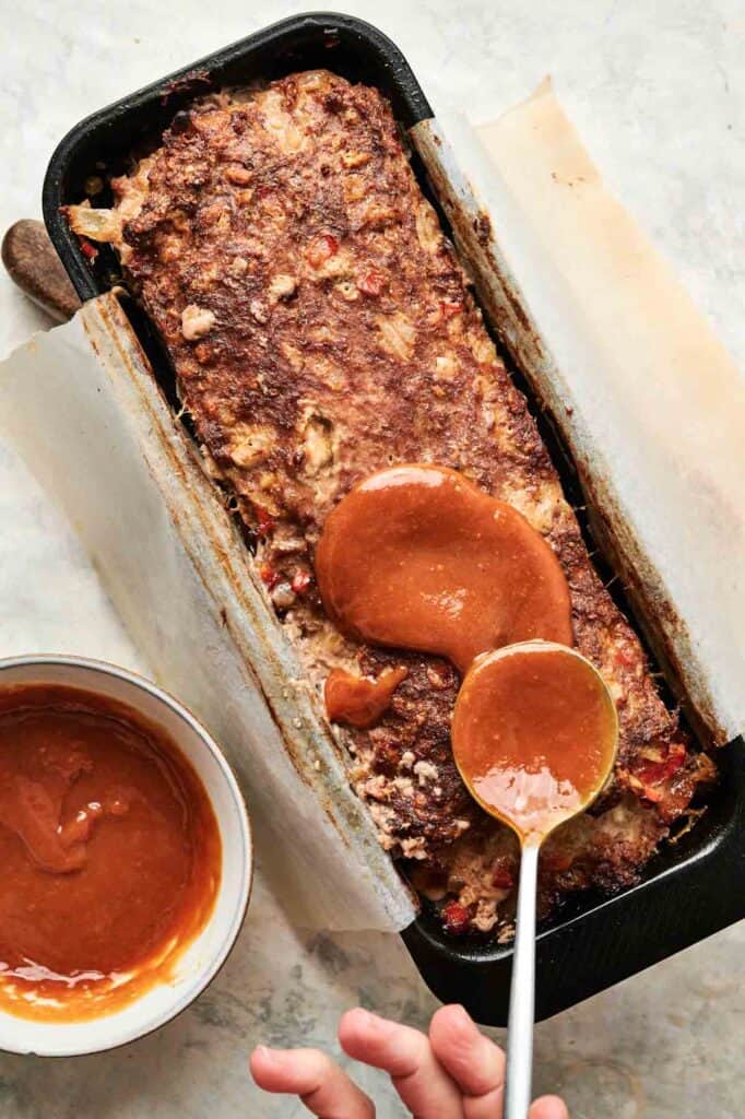 A hand spoons reddish-brown sauce onto a cooked Cracker Barrel Meatloaf in a parchment-lined loaf pan; a bowl of sauce sits nearby.