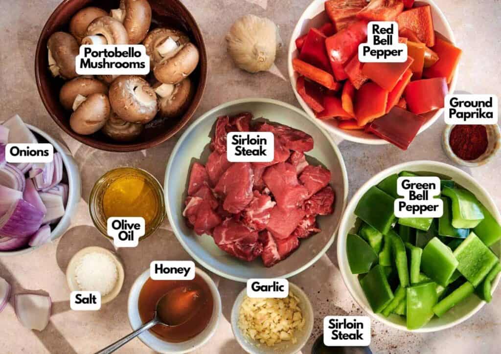 A top-down view of bowls containing ingredients for Grilled Steak Kabobs: portobello mushrooms, red and green bell peppers, sirloin steak pieces, onions, olive oil, salt, honey, garlic, and ground paprika.
