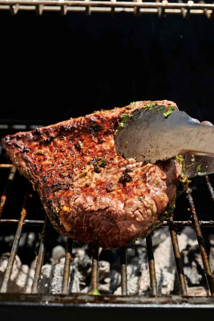 A large, seasoned grilled tri tip steak is being cooked over charcoal, with metal tongs holding one edge of the meat on the grill grate.