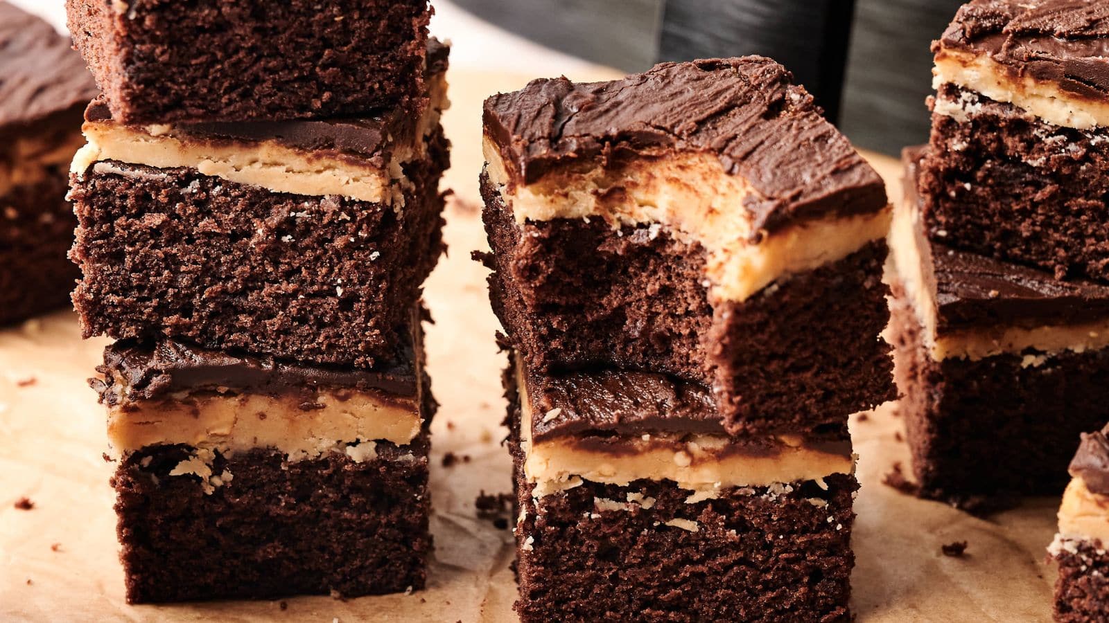 Several stacked chocolate brownies with a layer of peanut butter filling and chocolate frosting, placed on a parchment-covered surface.
