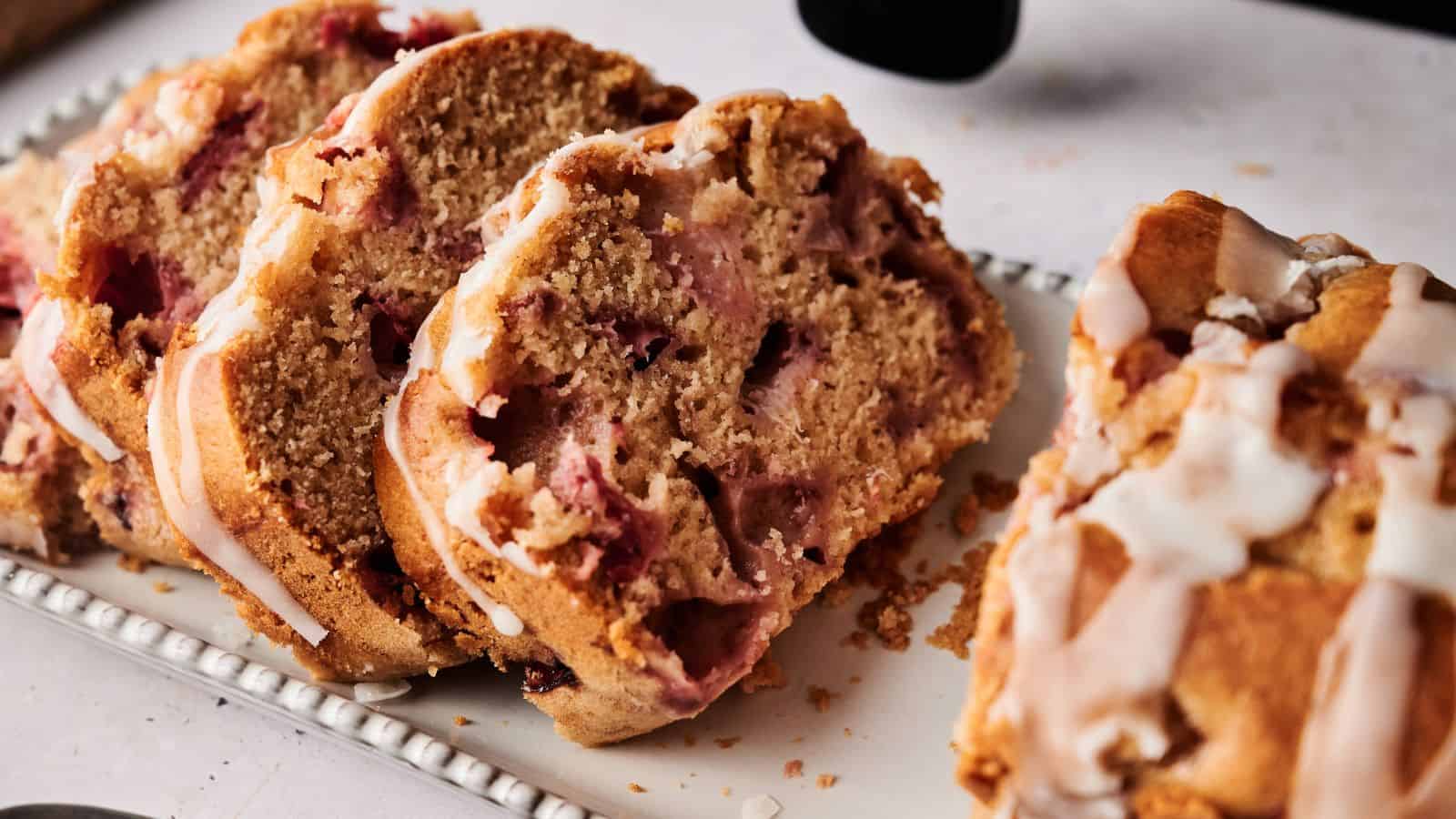 Three slices of strawberry bread with white icing are displayed on a rectangular plate, with the rest of the loaf nearby.