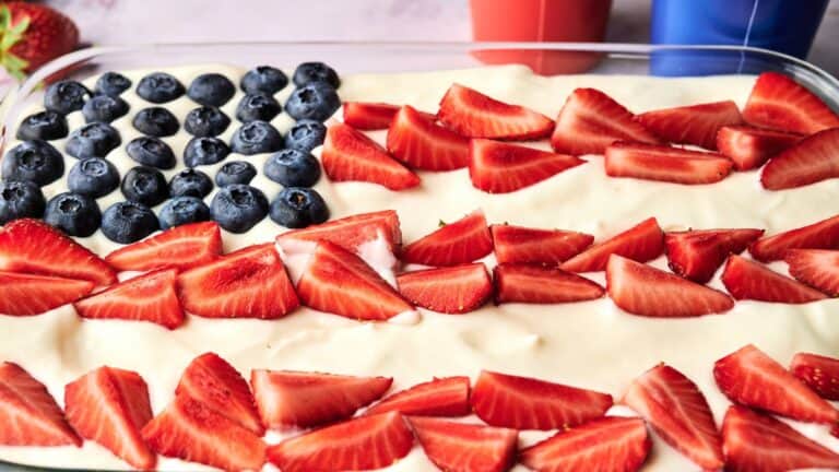 A rectangular dessert is topped with blueberries and sliced strawberries arranged to resemble the American flag on a creamy white layer.
