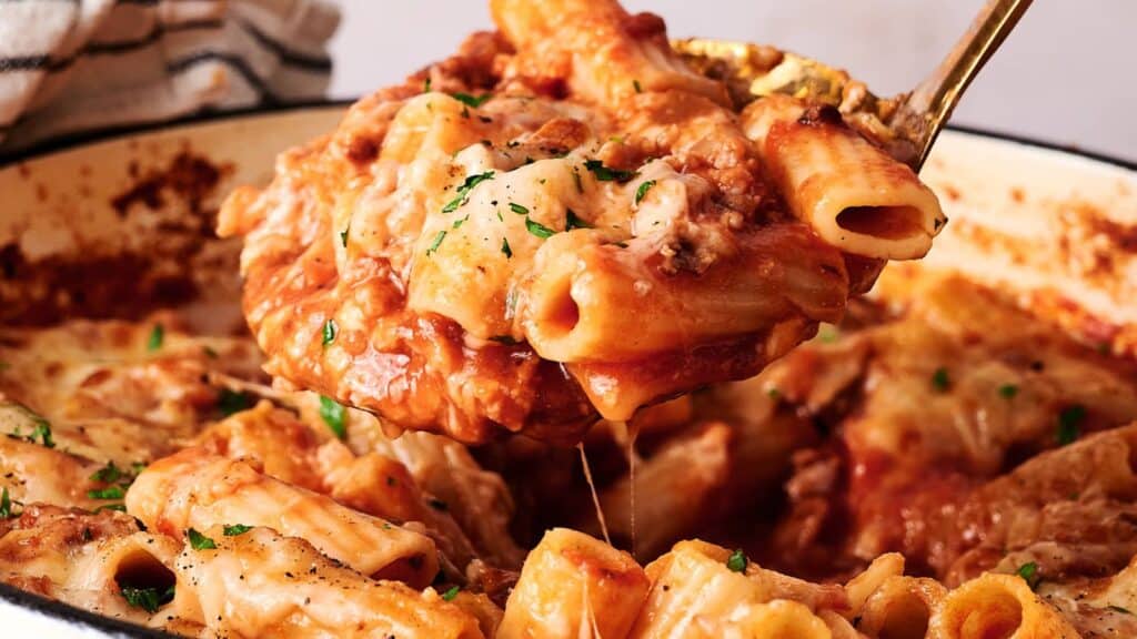 A close-up of baked ziti with melted cheese and tomato sauce being served from a casserole dish with a spoon.