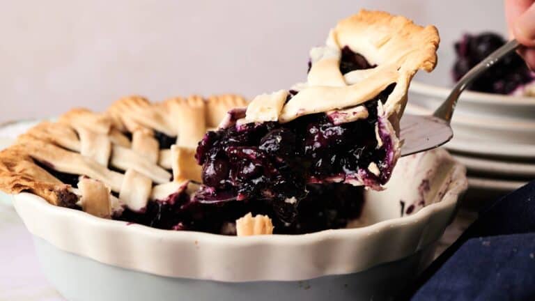 A slice of blueberry pie with a lattice crust is being lifted from a ceramic pie dish, showing the filling and flaky pastry.