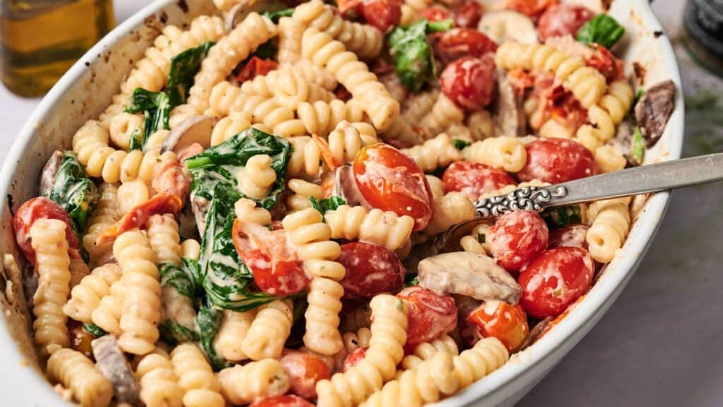 A ceramic dish filled with cooked curly pasta, cherry tomatoes, spinach, mushrooms, and a creamy sauce, with a serving spoon.