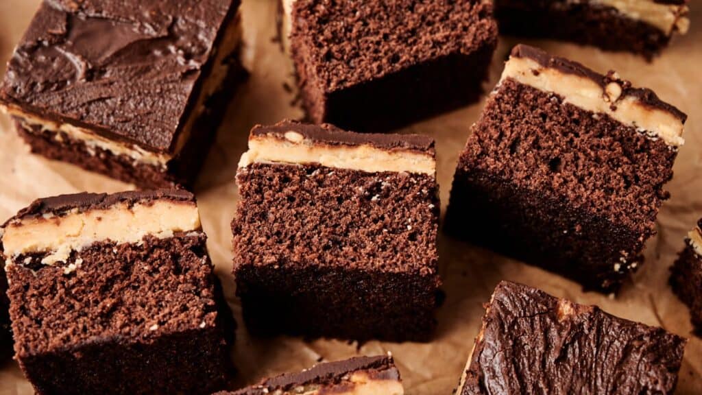 Several squares of chocolate cake with a layer of peanut butter filling and a chocolate topping are arranged on brown parchment paper.
