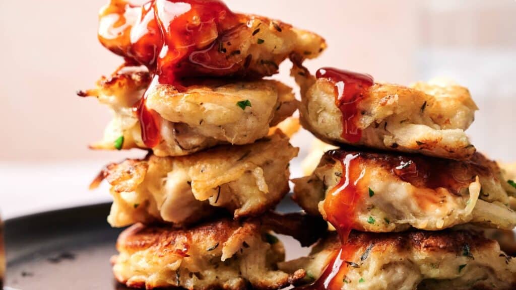 A stack of golden-brown fritters topped with a drizzle of red sauce, arranged on a dark plate.