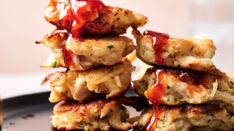 A stack of golden-brown fritters topped with a drizzle of red sauce, arranged on a dark plate.