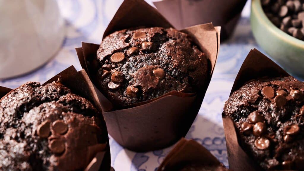 Close-up of chocolate muffins in brown paper wrappers, topped with chocolate chips, arranged on a patterned surface.