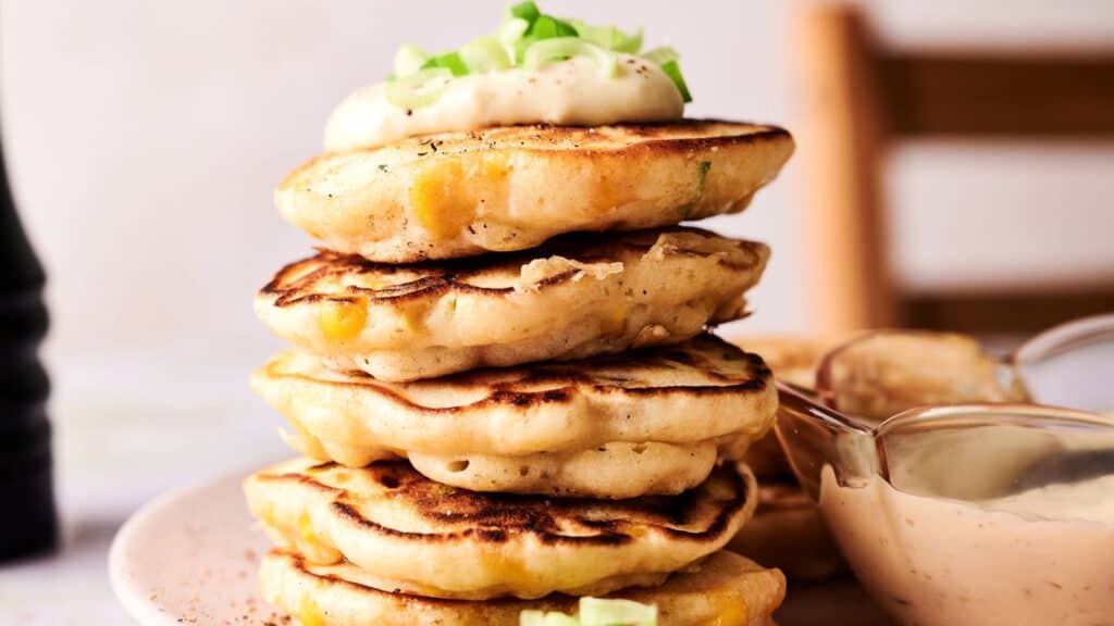 A stack of savory pancakes topped with a dollop of sauce and chopped green onions, with a bowl of dipping sauce beside them on a plate.