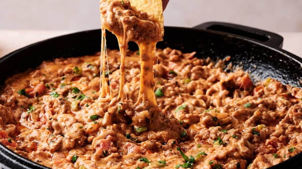 A close-up of cheesy ground beef dip in a skillet, with a tortilla chip being dipped and cheese stretching.